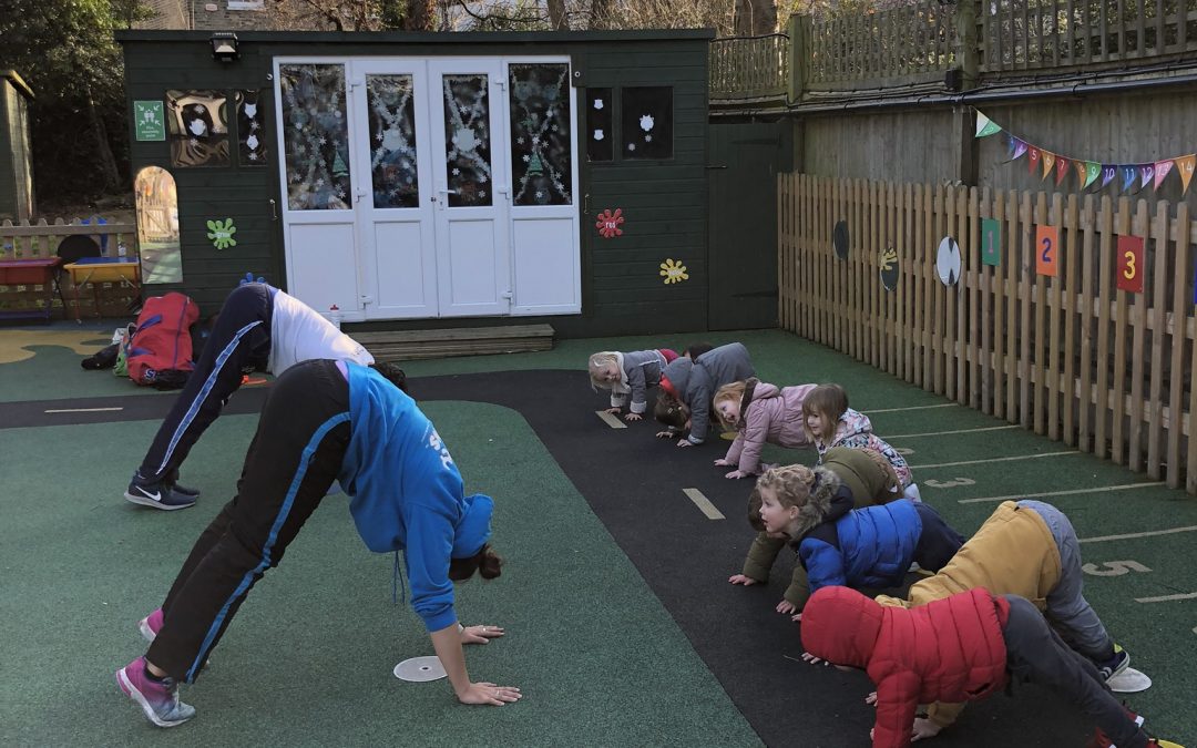 Two sports coaches leading a class of toddlers in yoga. The pose they are doing is downward dog.